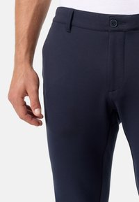 Pantalon bleu marine en tissu lisse et extensible. Il possède une fermeture à bouton et des poches avant. La ceinture est plate avec des coutures minimales.