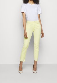 Jeans de mezclilla amarillos claros con un ajuste slim y dobladillo deshilachado, combinados con una camiseta de algodón blanca que presenta un pequeño detalle de bolsillo. Tacones altos nude.