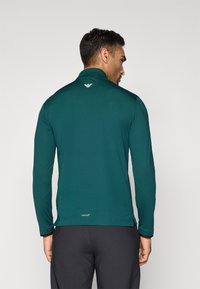 Muškarac u tamnozelenoj polosvijetloj košulji dugih rukava i crnim hlačama, okrenut leđima, s prikazanim logotipima na gornjem dijelu leđa i blizu struka.