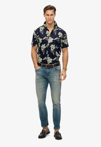 Camisa de manga corta azul marino con estampado floral, combinada con jeans azul claro y mocasines marrón oscuro. Cinturón y reloj marrón incluidos.
