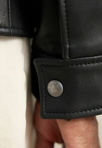 Schwarzes Lederjackenarm mit gestickten Details und einem silbernen Knopf mit der Aufschrift "SIGNATURE DURABLE GOODS". Leichte Stoffunterlage.