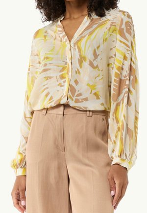 Woman wearing a long-sleeve blouse with yellow and beige abstract print tucked into high-waisted light brown pants.