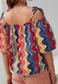 Femme portant un haut coloré à épaules dénudées avec des rayures verticales ondulées en rouge, bleu, jaune et vert, des bretelles nouées, et un pantalon rose.