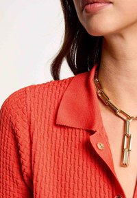 Czerwona dzianinowa koszulka polo z teksturowanym wzorem, wyposażona w kołnierzyk i złoty naszyjnik z ogniwami jako akcent.