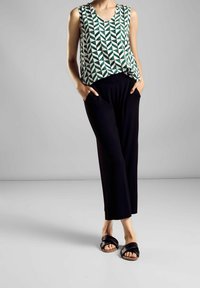 Ärmlös topp med grönt och vitt geometriskt mönster, kombinerad med svarta vida byxor. Modellen står med händerna i fickorna och bär svarta sandaler.