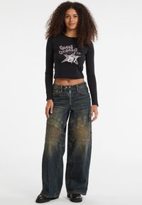 Schwarzes, langärmliges, gekürztes Oberteil mit "Guess Originals" Grafik; weiter Beinen Jeans aus blauem Denim mit strukturierten Patchwork-Details und einem ausgewaschenen Finish.