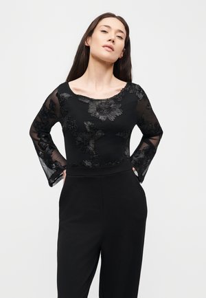 Une femme pose les mains sur les hanches, vêtue d’une combinaison noire aux manches transparentes en dentelle florale et au corsage orné.