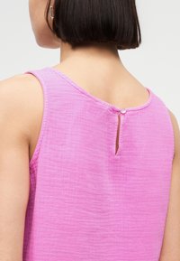 Femme aux cheveux courts et foncés portant un haut rose texturé sans manches avec une petite fermeture à bouton à l'arrière de l'encolure.