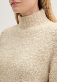 Beżowy sweter z wysokim kołnierzem wykonany z fakturowanego, puszystego materiału. Tkanina wydaje się miękka i ciepła, z lekko luźnym krojem i minimalnymi detalami.