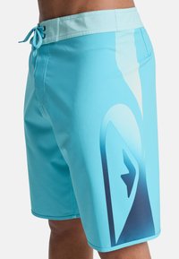 Ανοιχτό μπλε μαγιό σορτς κατασκευασμένο από απαλό ύφασμα, με μοτίβο διαβάθμισης σε πιο σκούρες μπλε αποχρώσεις και ελαστική μέση με κορδόνι.