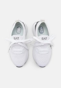 Valkoiset urheilukengät, joissa on teksturoitu neulottu päällinen, litteät nauhat ja logon yksityiskohdat kielekkeessä ja kannassa. Pyöristetty pohja, jossa kontrastievä musta ulkopohja.