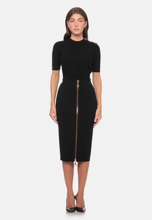 Donna in piedi che indossa un vestito nero aderente con maniche corte e una cerniera dorata frontale che va dalla vita all'orlo, abbinato a tacchi neri.