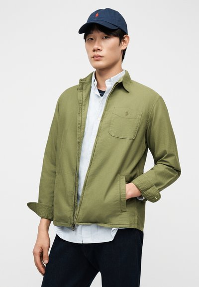 Νέος άνδρας που φοράει πράσινο μπουφάν με φερμουάρ πάνω από ριγέ πουκάμισο, μπλε παντελόνι και μπλε καπέλο μπέιζμπολ με κόκκινο λογότυπο, στέκεται με το ένα χέρι στην τσέπη.