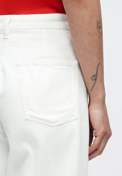 Λευκό παντελόνι από τζιν με πίσω τσέπη που διαθέτει διακριτική λεπτομέρεια ραφής. Ορατή λεία υφή, συνδυασμένη με κόκκινο τοπ που φαίνεται μερικώς.