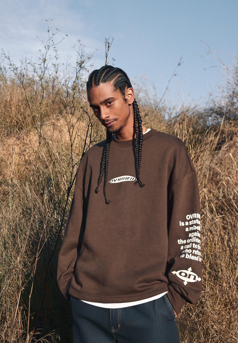 Smešani dukserica u opuštenom kroju s bijelim natpisom na desnoj rukavu i logom s lijeve strane, uparena s tamnim trapericama.