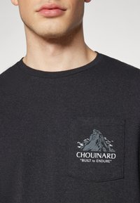 T-shirt en coton noir avec un col rond, présentant une illustration de poche sur la poitrine d'une montagne et le texte "Chouinard Built to Endure."