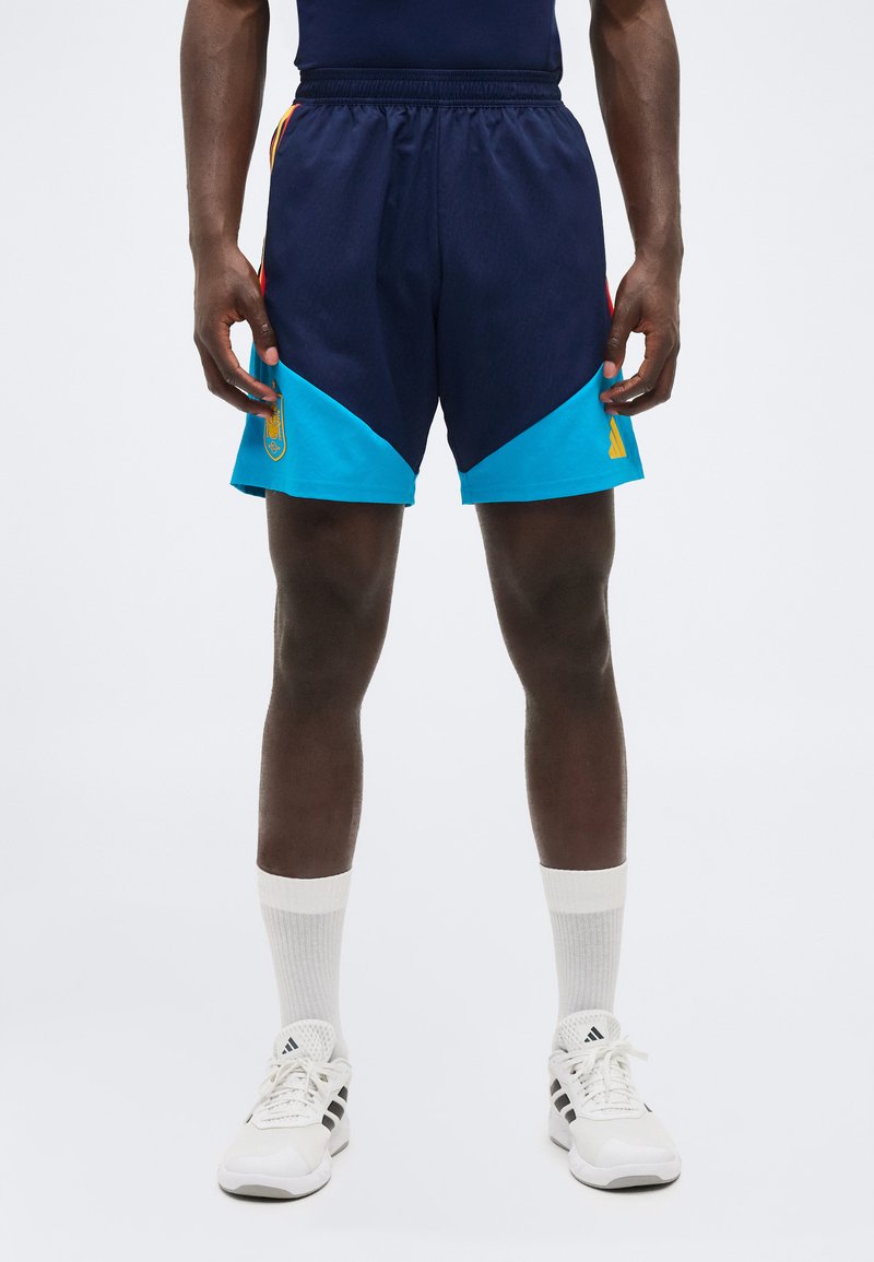 Άνδρας που φοράει σπορ σορτς σε ναυτικό και ανοιχτό μπλε χρώμα, στέκεται, με λευκά παπούτσια Adidas και κάλτσες, σε μονόχρωμο φόντο.