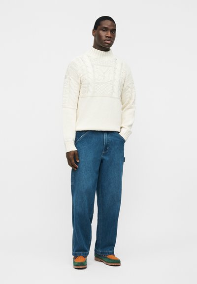 Νεαρός άνδρας στέκεται με τα χέρια στις τσέπες, φοράει λευκό πλεκτό πουλόβερ με υφή, φαρδιά μπλε τζιν και δίχρωμα καφέ και πράσινα παπούτσια.