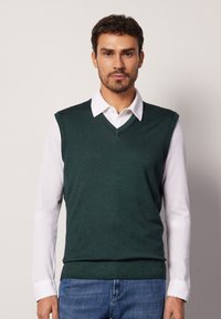 Zielony dzianinowy kamizelka w kształcie litery V, noszona na białej koszuli z kołnierzykiem. Posiada prążkowany dół i otwory na ramiona; zestawiona z niebieskimi dżinsami.