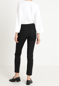 Bílá dlouhá halenka s elastickým pasem v kombinaci s černými slim-fit kalhotami. Černé boty doplňují outfit. Zobrazený zadní pohled.