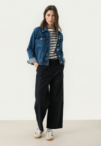 Denim jacket with button closure, medium blue color, worn over a black-and-white striped shirt. Black wide-leg trousers and white sneakers complete the look.