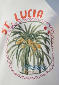 Valkoinen kangas, jossa on brodeerattu ympyräkuvio, jossa on vihreä palmupuu, oransseja yksityiskohtia, sininen aalto ja punaisia koriste-elementtejä. Tekstissä lukee "ST. LUCIA."