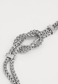 Bracelet en métal argenté avec des maillons entrelacés, présentant deux accents gravés "GUESS". Texture lisse avec une finition polie.