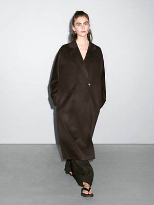 Long, dark brown coat made of a smooth material, featuring a double-breasted design, large lapels, and side pockets. Worn over wide-leg trousers.