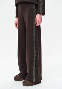 Bruine wijde broek met een gladde textuur, voorzien van contrasterende decoratieve biezen langs de zijkanten. Draagt donkere bruine pantoffels.