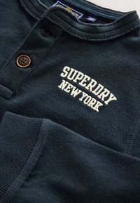 Mörkgrön bomulls-långärmad tröja med henley-krage, med en brun knapp och "SUPERDRY NEW YORK" broderad i vitt.