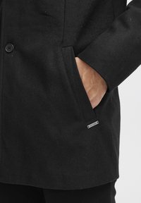 Abrigo de mezcla de lana negro con una textura suave, que presenta dos botones y bolsillos laterales. Una pequeña etiqueta de metal es visible cerca del bolsillo.