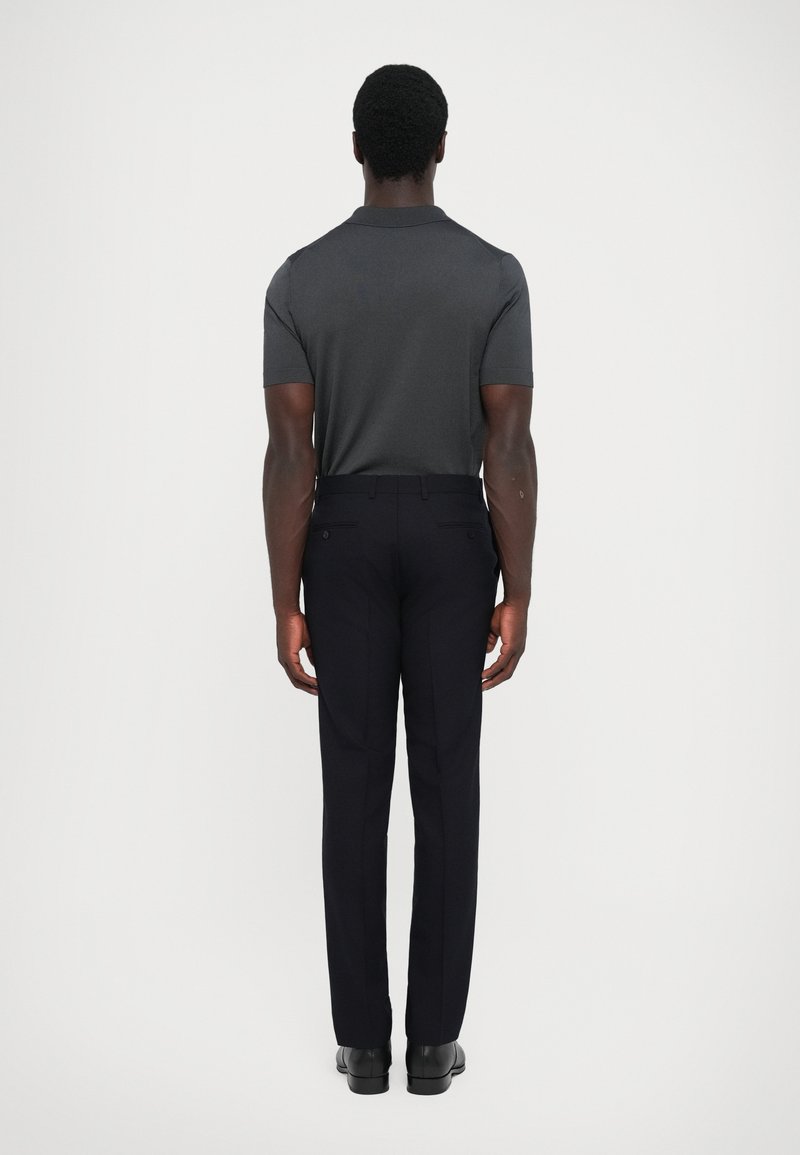 Kurzärmliges, dunkelgraues Poloshirt, kombiniert mit maßgeschneiderten, marineblauen Hosen. Die Rückansicht zeigt einen glatten Stoff und ein minimalistisches Design. Schwarze Schuhe.