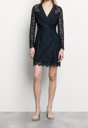 Kobieta ubrana w czarną, długorękawową koronkową sukienkę w kwiaty z kopertowym dekoltem, w towarzystwie srebrnych sandałów na obcasie z paskiem wokół kostki, stojąca na jednolitym tle.