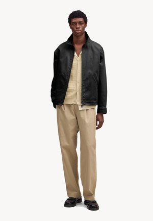 Jonge man die een zwart vest met rits draagt, een beige gebreid vest, lichtbeige broek en zwarte loafers, staand tegen een effen achtergrond.
