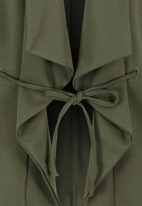 Tissu vert olive avec de larges cols noués en un nœud lâche au centre, montrant des coutures détaillées et des couches pliées.