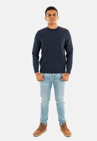 Maglione a maglia blu navy con scollo a girocollo, caratterizzato da quattro bottoni sulla spalla sinistra. Indossato con jeans azzurri chiari e stivali beige.
