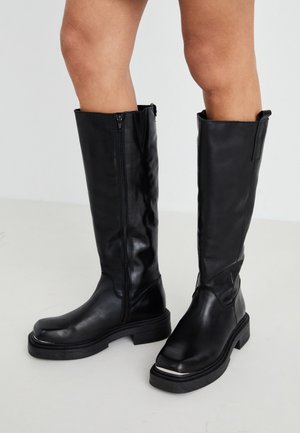 Põlve kõrgused mustad nahast saapad, millel on külgutte ja paks kummist tallad, ning tekstuuriga viimistlus minimalistliku disainiga.