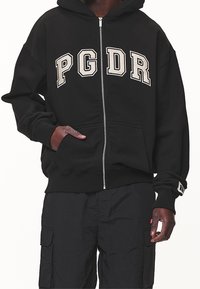 Černá mikina na zip s velkým krémovým logem "PGDR" na hrudi, s přední kapsou a ribbovanými manžetami.