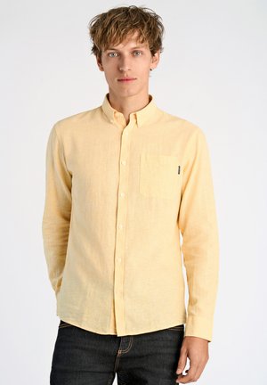 Camisa de botones de manga larga color amarillo claro en una mezcla de algodón texturizado, con un bolsillo en el pecho y cuello clásico. Combinada con jeans oscuros.