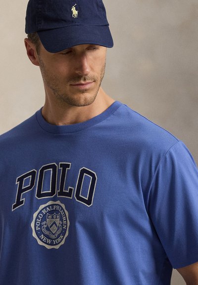 Άνδρας που φοράει μπλε μπλουζάκι Polo Ralph Lauren και σκούφο σκούρου μπλε με λευκό κεντημένο λογότυπο, κοιτάζει προς τα κάτω πλάγια.