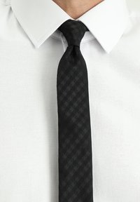 Cravatta nera a motivo geometrico realizzata in tessuto lucido, indossata con una camicia bianca con colletto.