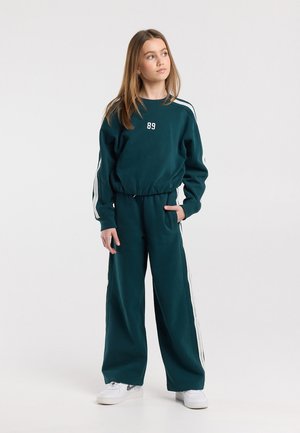 Ragazza in piedi con la mano in tasca, indossa una tuta verde scuro con strisce bianche e il numero "89" sul petto, sneakers bianche, guarda di lato.