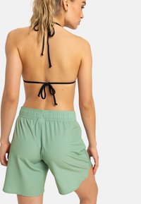 Čierny halter bikiny top s viazaním na chrbte, v kombinácii so svetlozelenými plavkovými šortkami po stehná, ktoré majú elastický pás a zakrivený lem.