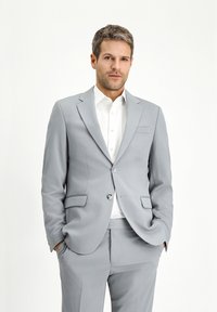 Hellgrauer Anzug mit Reverskragen, einreihiger Knopfverschluss und aufgesetzten Taschen. Darunter ein weißes Hemd, glattes Stoffgefühl.