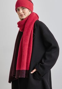 Gorro de punto rojo y bufanda a juego, textura suave con un color degradado de rojo a burdeos oscuro, llevados sobre un abrigo de lana negra con un bolsillo.