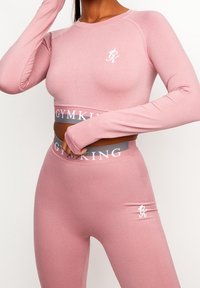 Růžový dlouhý rukáv crop top a odpovídající legíny s přiléhavým siluetou. Obsahuje akcenty loga a kontrastní pás s názvem značky.