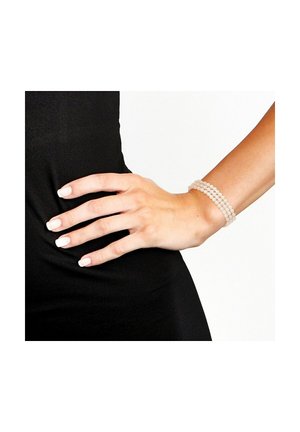 Bracelet extensible composé de petites perles beige translucides. La main a un vernis à ongles neutre et repose sur une surface en tissu noir.