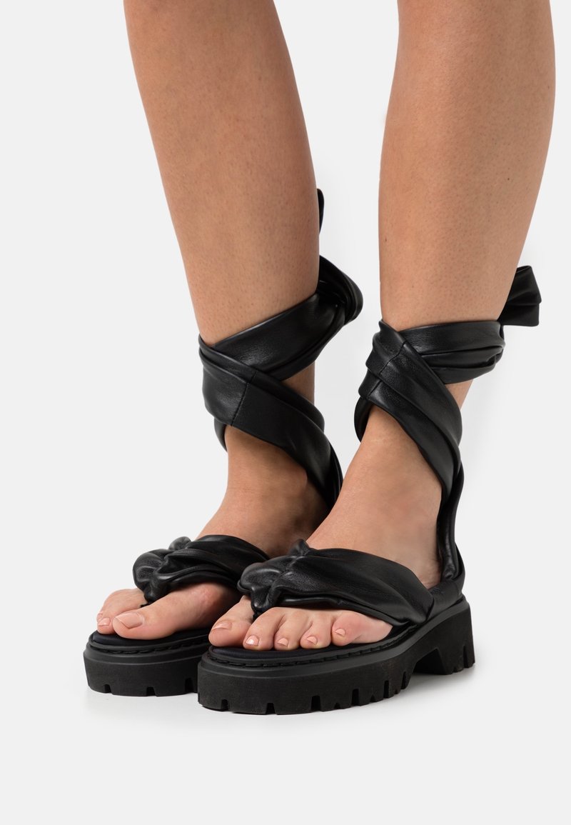 Czarne skórzane sandały z skręconymi ramiączkami wokół kostek, masywnymi czarnymi podeszwami platformowymi i otwartym noskiem. Wyróżniający się teksturowany detal skórzany.