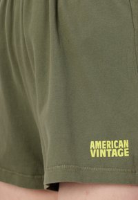 Zelené bavlnené šortky s voľným strihom. Obsahujú žlté logo "AMERICAN VINTAGE" a viditeľný šev pozdĺž lemu.
