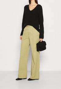 Schwarzer Strickpullover mit V-Ausschnitt. Hellgrüne weit geschnittene Hose. Eine schwarze strukturierte Handtasche und schwarze spitze Ballerinas runden das Outfit ab.